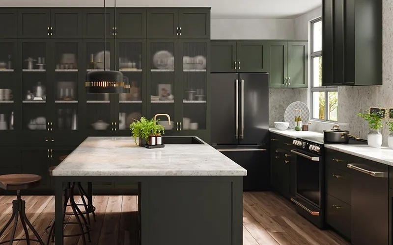 A modern kitchen with dark green cabinets, brass hardware, and a large light gray marble island. The space features a black refrigerator, black oven, open glass-front cabinets, and small potted herbs on the countertop