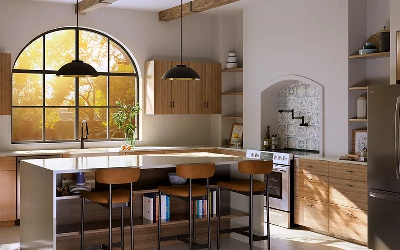 A warm kitchen lit by sunlight streaming through a large arched window. The space has light wood cabinets, a white island with open shelving, brown bar stools, and decorative tile behind the stove