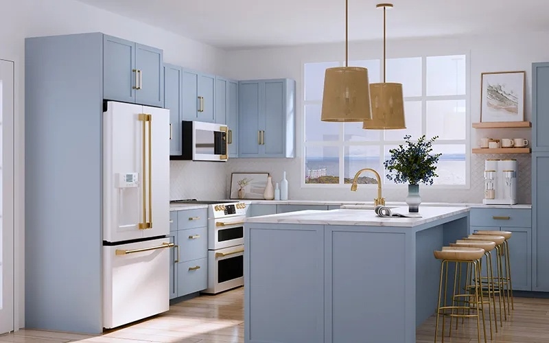 A bright kitchen with light blue cabinets, white countertops, and brass accents. The room includes a white refrigerator, double oven, woven pendant lights, and a window overlooking a coastal view